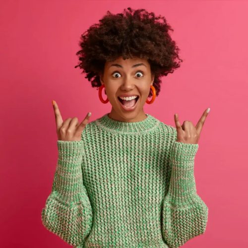 happy-carefree-dark-skinned-rebellious-young-woman-enjoys-awesome-music-makes-rock-n-roll-gesture-has-fun-on-music-festival-or-cool-event-wears-casual-jumper-poses-against-pink-wall